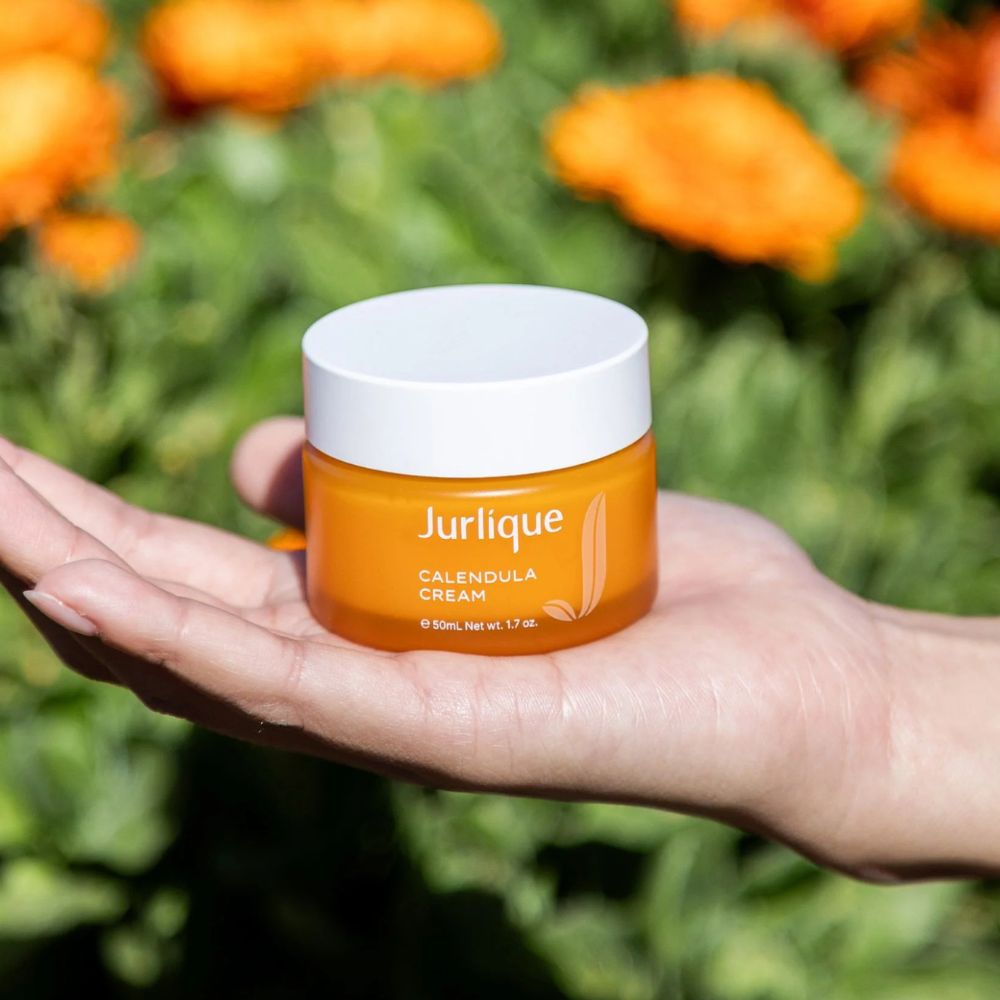 Hand holding a jar of Jurlique Calendula Cream with flowers in the background